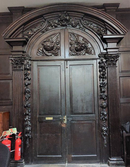 Dark wood double doors with elaborate carved archway above, with carved columns to either side, with garlands of fruit and flowers. Between doors and arch are another decorated band, and two quarter round sections with cherubs/angels.
To the left are two red fire extinguishers.