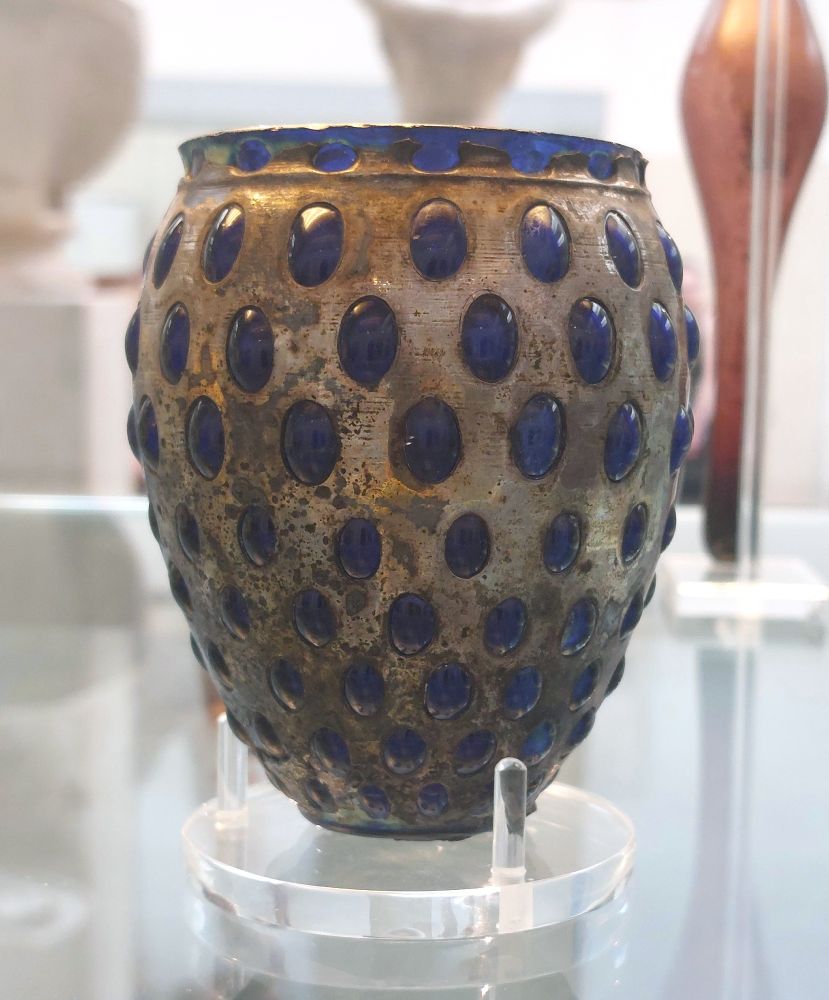 A bulbous vessel, with a smaller base and curving in slightly to rim at top. It is cased in silver, with oval shapes cut out all round, larger at the wider section than at the more narrow bottom and top. Deep blue glass has been blown into the casing so that cushions of blue glass protrude out through the ovals.