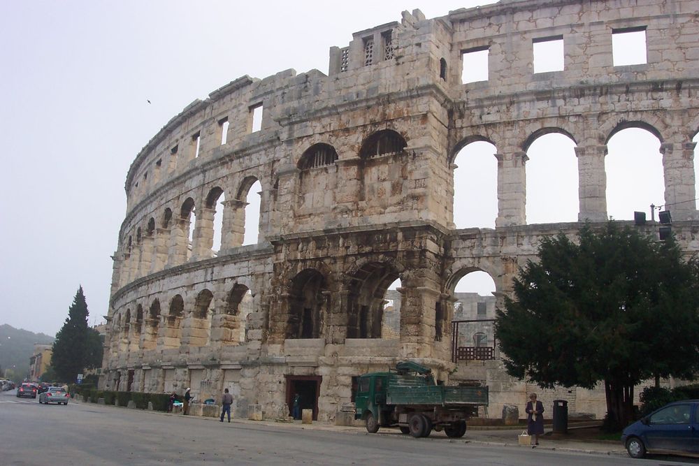 External view of substantial white stone amphitheatre with arches of three storeys visible with a substantial tower with 2 arches on 1st and 2nd levels - these held cisterns for spraying water on audience. There is also a lower level visible, without arches.
The roadway outside has a few cars and trees alonside the building.