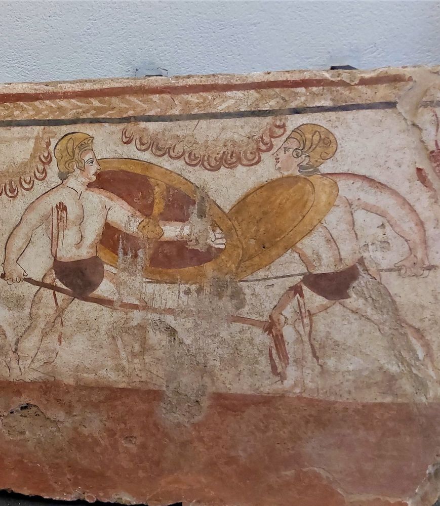 Two warriors clash with shields and spears, shoeless and wearing only briefs and helmets. One on the left has bleeding wound in his chest, one on left has one on his left thigh. 
Colours are a deep red and gold on white background, and the figures have shading and lines to emphasise their strong physiques. 
The ground is a lighter red and there is a band with black and dark red, top and bottom of a light red continuous arrow shaped motif.
