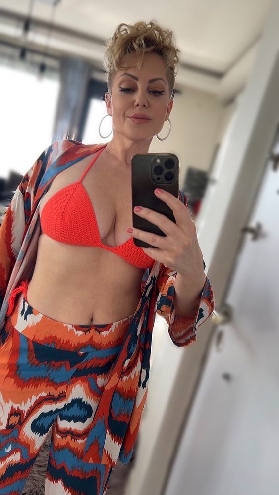 A stunning mirror selfie of a blonde woman with short pixie hair and radiant green eyes, capturing her soft beauty and confident aura. She’s wearing a red bikini top under a light beach cover-up, the delicate straps hugging her chest and showing off her natural curves without revealing too much. Her toned stomach peeks out, and the open front of the outfit frames her upper body like a tease. Her gaze is calm and inviting, effortlessly seductive in a way that feels both classy and dangerously tempting. With sunlight kissing her skin and beachy tones in the background, this photo feels like a private moment stolen between ocean breeze and mischief. Tags: #blonde #mirrorpic #bikinigirl #beachlook #onlyfans