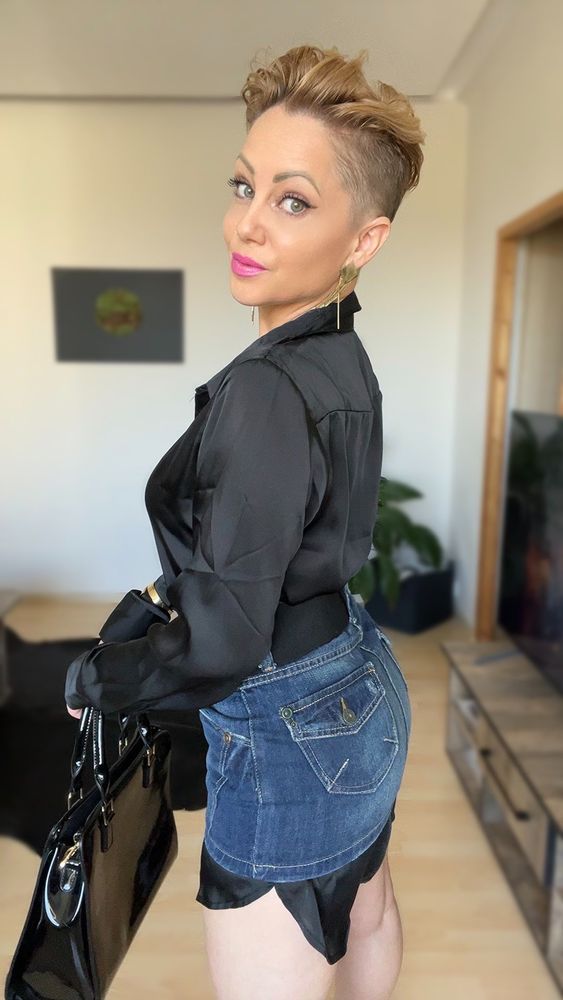 Full-body photo of a breathtaking blonde woman with striking green eyes and a short, edgy pixie haircut—cleanly shaved on the sides and left longer on top, giving her a bold yet feminine look. She stands confidently outdoors, one hand resting near her hip and the other gently gripping a sleek black shoulder bag. She wears a fitted black shirt that hugs her waist and a short denim mini skirt that shows off her sculpted legs. The lighting highlights the glow of her skin and the way the fabric moves with her posture. Her expression is effortless but piercing, a quiet kind of power. Her stance is casual, yet undeniably commanding—one hip cocked just slightly, hinting at the curve of her body beneath the clothes. The skirt rides up just enough to tease, but not enough to reveal. Every detail from her confident eyes to her subtle smirk to the way her shirt tucks into the waistband feels intentional, captivating. A moment frozen in beauty, attitude, and quiet seduction.