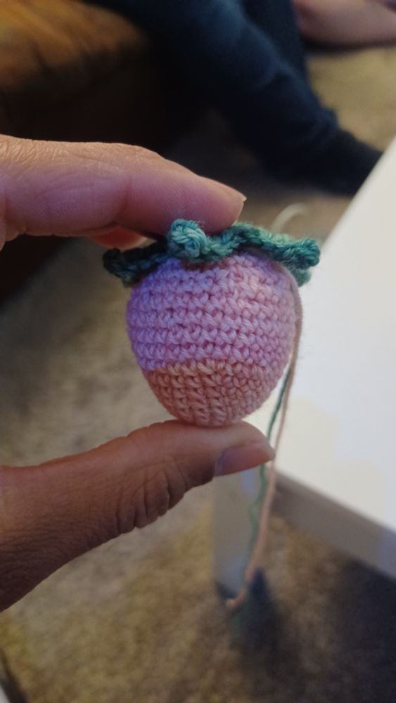 A hand holds up a single crochet amigurumi pink strawberry, now with the green crochet leaves attached. 
