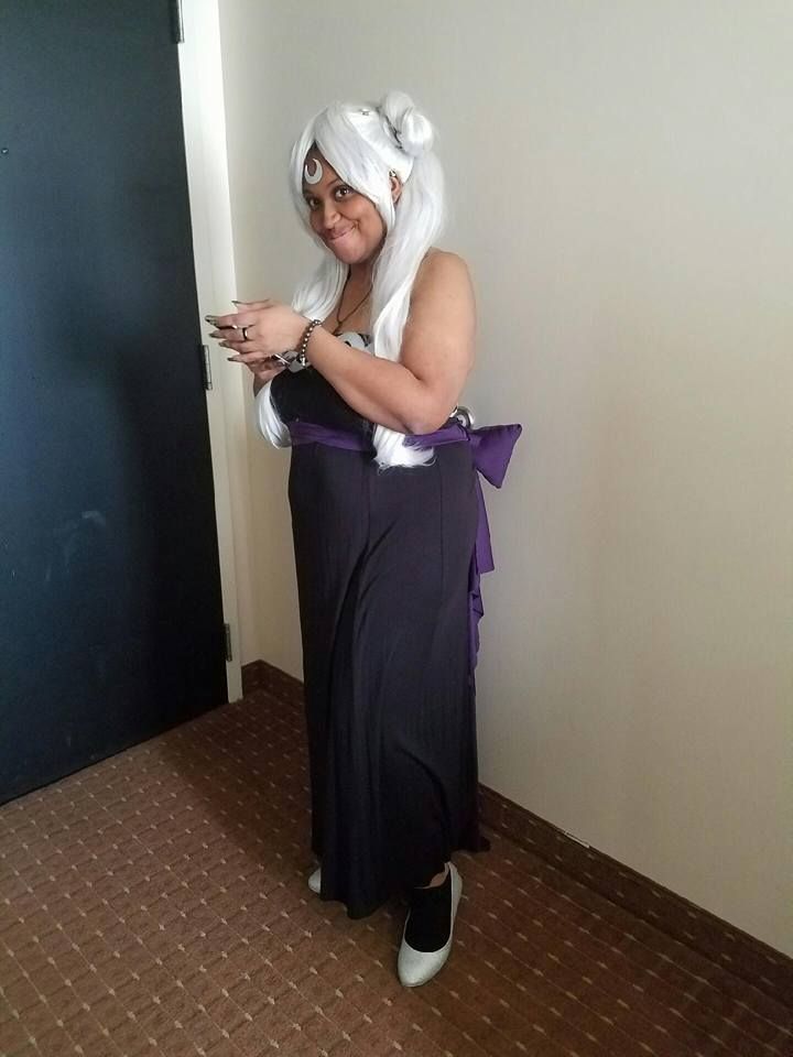  Christina cosplaying as an alternate universe dark Princess Serenity from Sailor moon. She is wearing a black gown with a purple bow. On her feet are silver shoes and on her head is a silver wig styled with a bun and ponytail on each side as well as a silver crescent moon on her forehead.