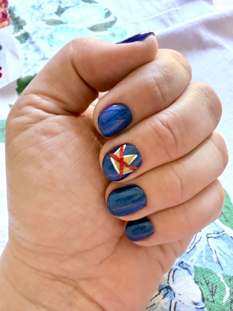 Left hand of a white person with short, blue-painted nails and a yellow, white and red No Kings crossed-out crown on the middle finger