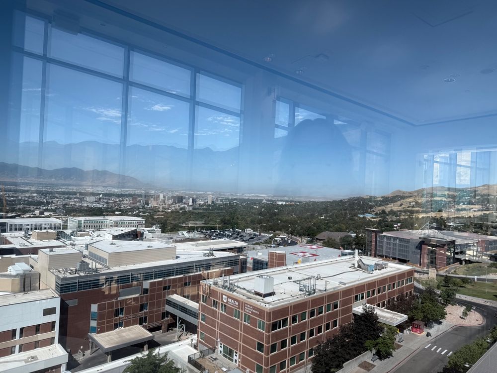 View from the 6th floor of Huntsman Cancer Center in Salt Lake City showing hospital buildings, the downtown area, and haze over the Great Salt Lake under a deep blue sky. The woman taking the photo is reflected in silhouette