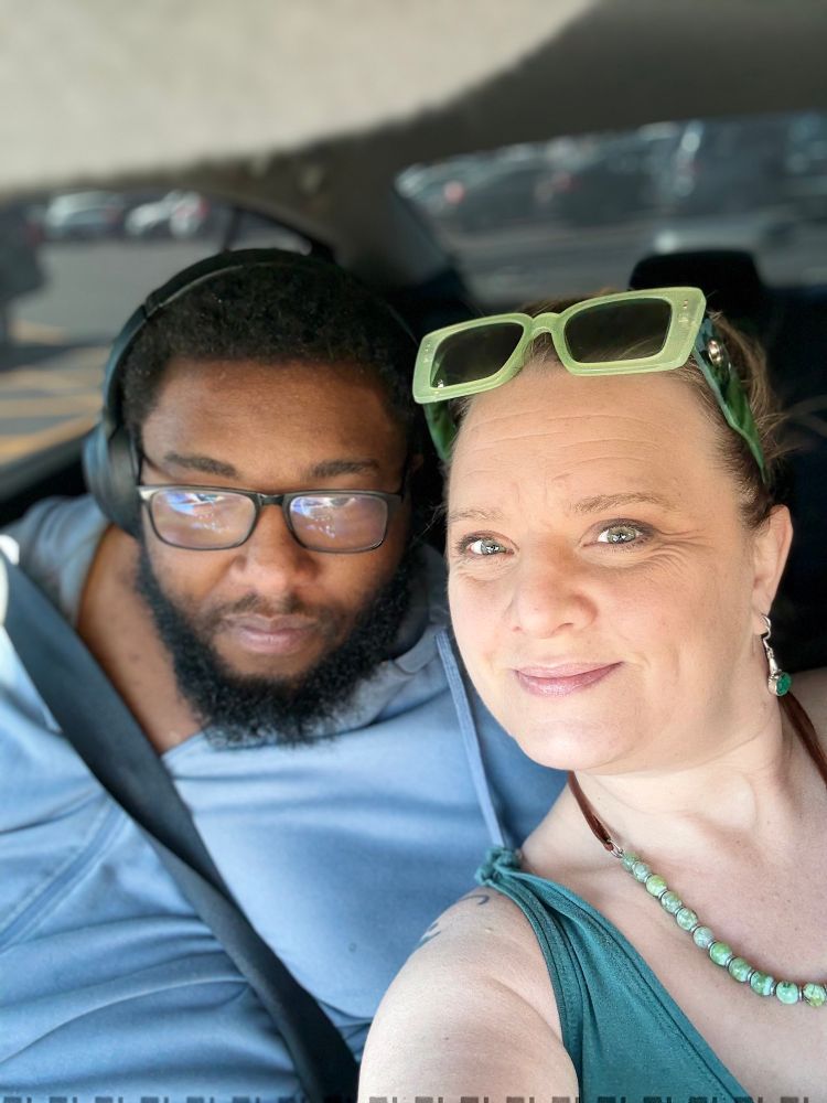 Mother and son, a white woman in her 50s and a Black man in his 20s, smile for a selfie inside a car