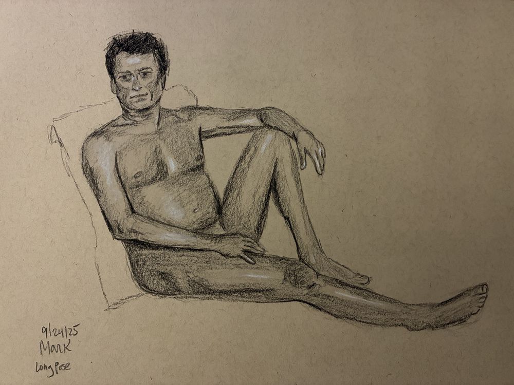 A nude male model sits on the floor with his back against the seat of an armchair. His right leg is straight before him, and his left knee is raised. His left forearm rests on his left knee, and his left hand is on his left thigh.