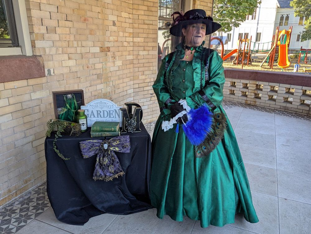 Madame Dutra stands in Victorian garb while standing next to her table of potions, books, and deadly plant lore.