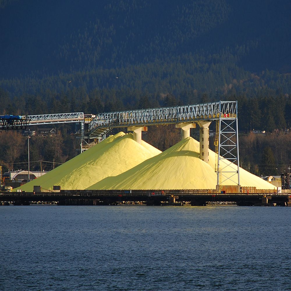 Photo of two warehouse-sized piles of bright yellow sulphur. Water in foreground leading up to industrial dock, trees going up mountain in background.