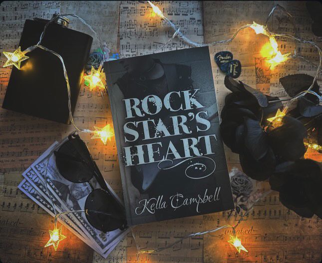 A book portrait: a paperback edition of Rock Star’s Heart rests on a background of sheet music with star-shaped lights, guitar picks, sunglasses, money, and black roses.