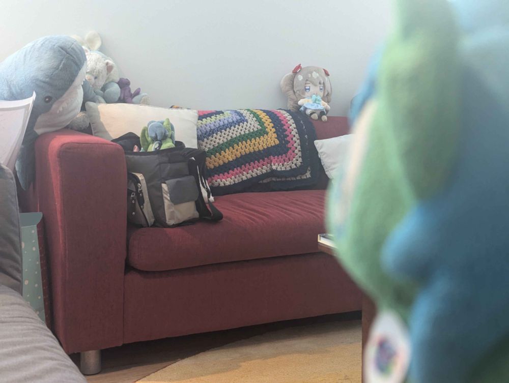 A photo of a red sofa with a bag that has the Old Alope Plush inside peeking out. On the right, next to us, is the Smol Alope plush looking at the Old Alope Plush. Also on the red sofa is a Blahaj shark, a plush bunny, a small plush purple teddy bear, and a plush of Neuro-Sama. 