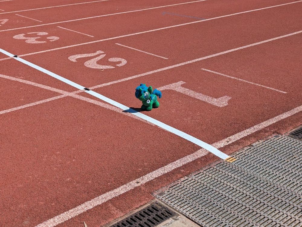 The Smol Alope Plush sits at the starting line of a big bright red athletic track at the number 1 position, ready to do a big run. It's quite sunny outside.