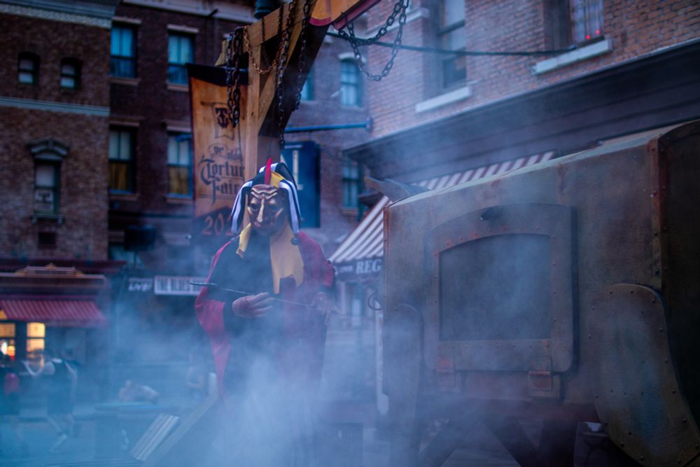 A jester stands next to a sculpted metal bull with smoke pouring out of it.