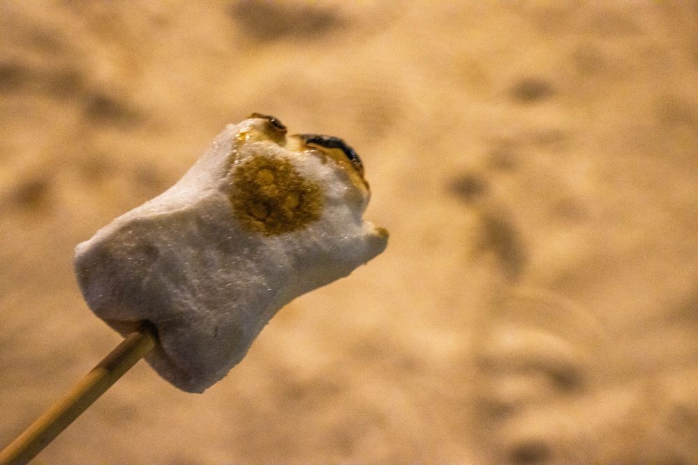 A ghost-shaped marshmallow, with its face golden brown, on a stick above the sand.