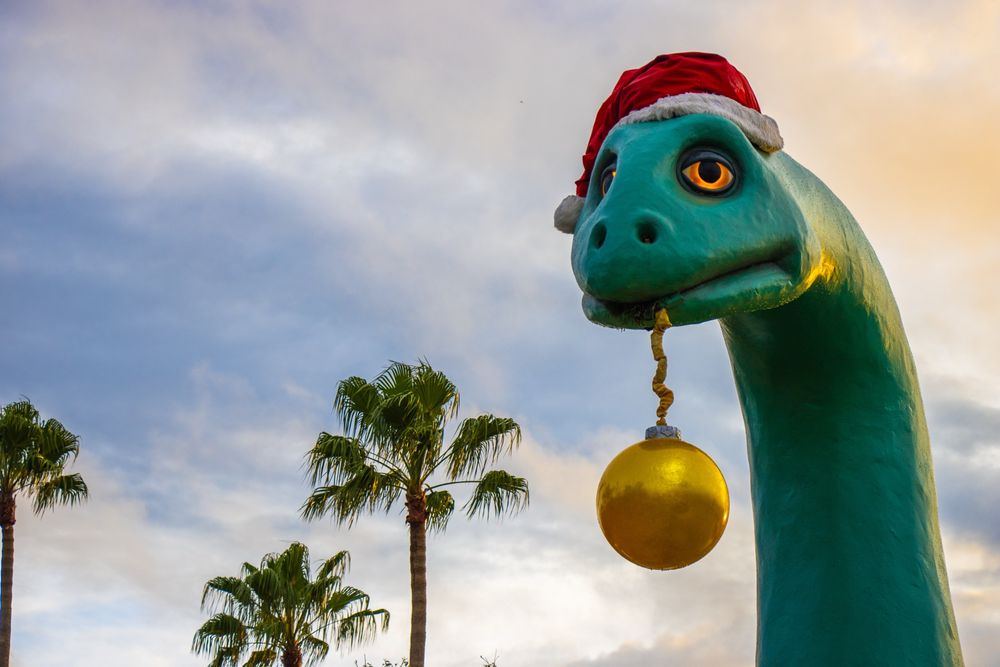 Gertie the dinosaur, wearing a Santa hat, holding a golden Christmas bauble in her mouth, at sunset with palm trees in the background.