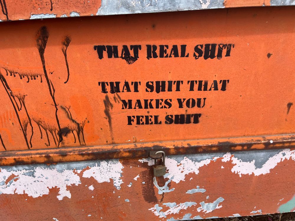 Black spray paint stencil on an orange beam that says “That real shit, that shit that makes you feel shit”