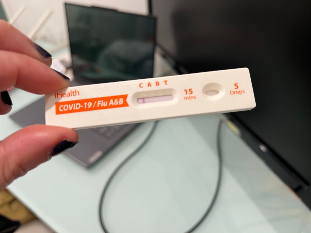 A hand holding a white and orange Covid test. The sample window says CABT to cover the control sample, flu A, flu B, and the test sample for Covid. Only the control sample line is highlighted. 