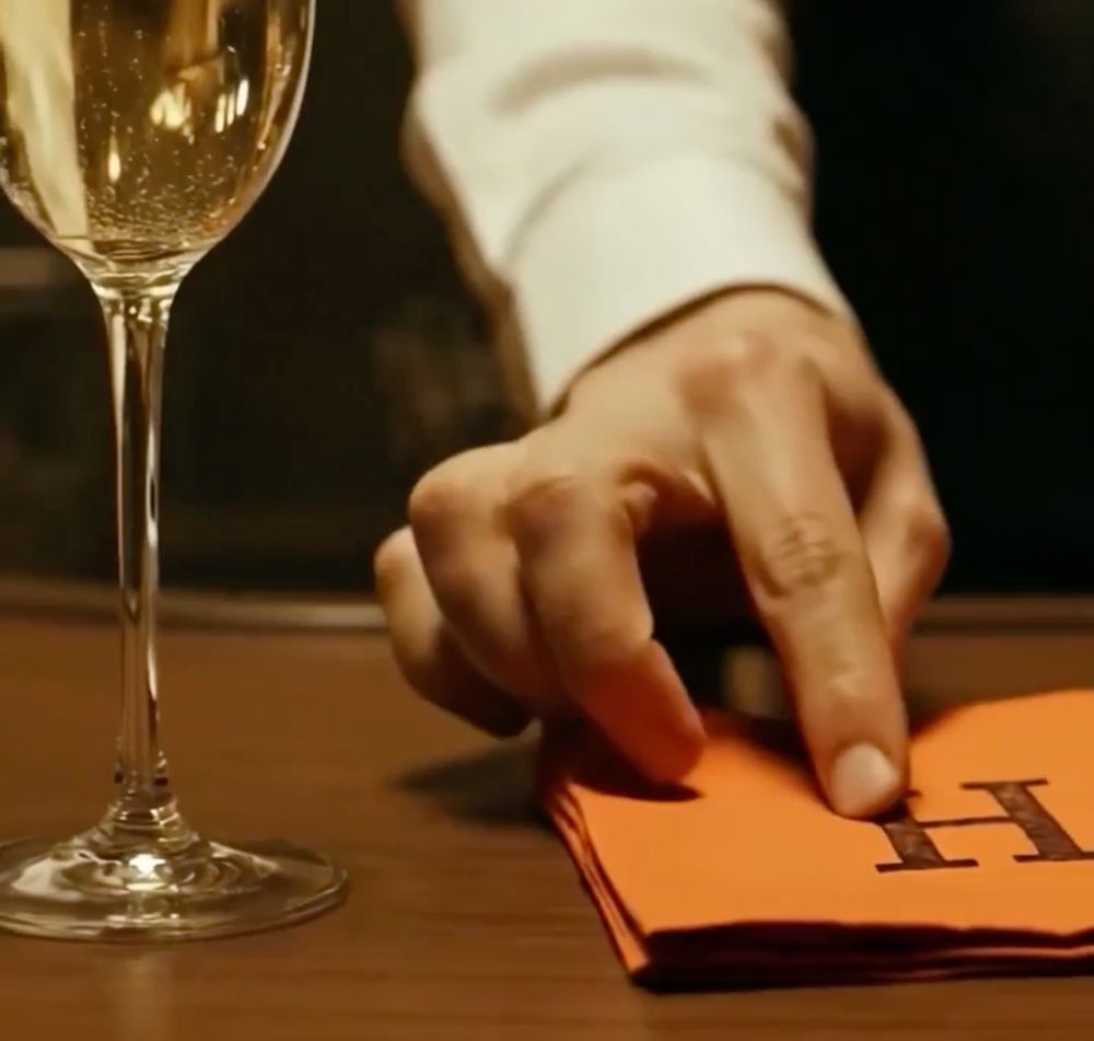 Screenshot of Taylor Swift AI promo video.

A young white man's hand rests on top of a thick orange square paper napkin with a black capital H printed in the middle. Next to it is a flute glass filled with champagne on a medium wood bar counter. The man is the bartender and he is in the act of giving us the napkin and sliding it towards us. His visible forearm is wearing a formal white button down shirt. However his extended pointer finger has a deformed third knuckle that is off center.