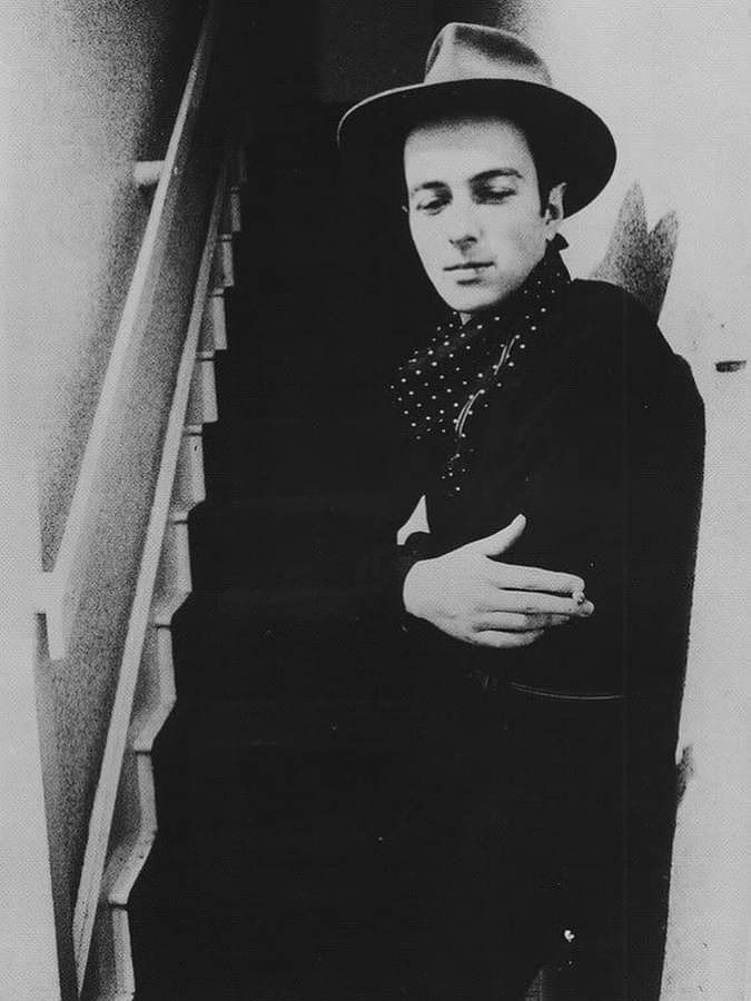 Young Joe Strummer in black, wearing a fedora and a scarf, smoking and looking introspective, dreamy, and stoned