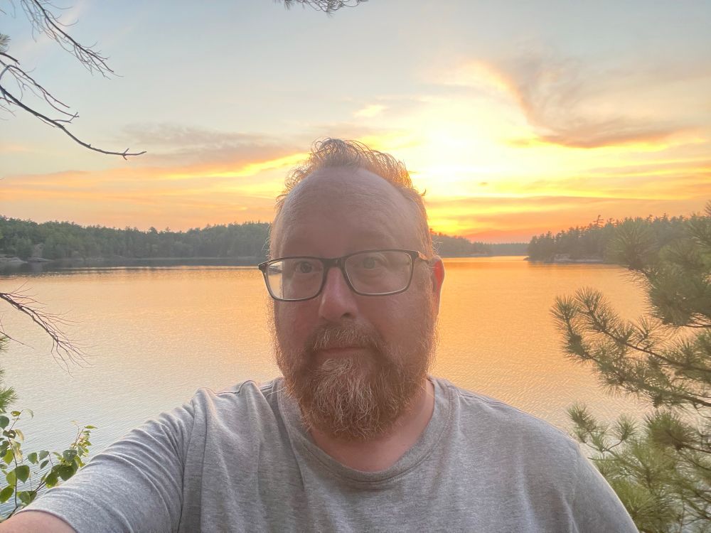 Selfie of me wearing a grey T-shirt with the sunset behind me. 