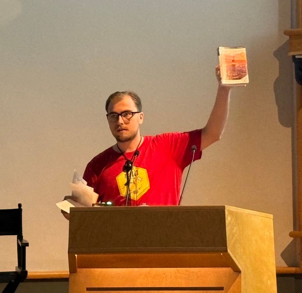 Speaker at podium holding book in one hand and loose pages in another 