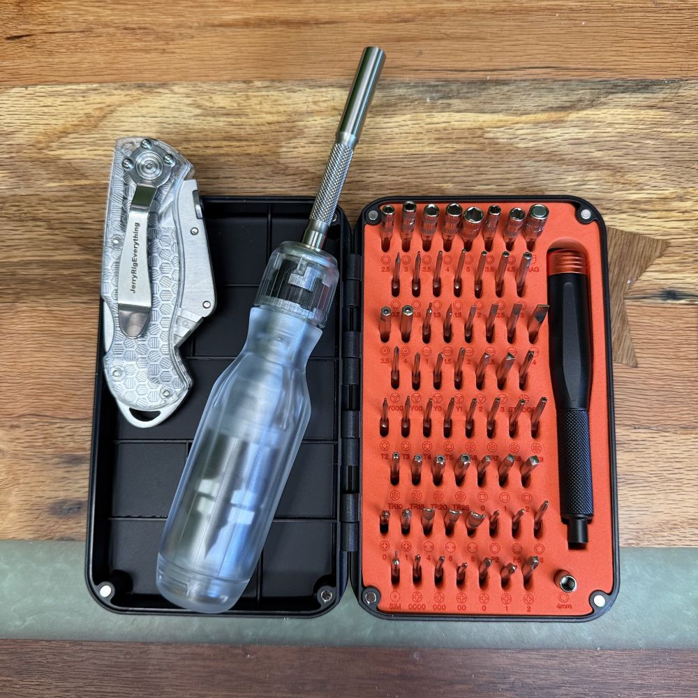 Photo tools sitting on my desk (horizontal slats of spalted oak, greenish resin, and a darker line near the bottom.

JerryRigsEverything knife: folding knife that uses utility knife blades. The handle is transparent plastic with a hexagonal pattern.

Linus Tech Tips ratcheting screwdriver, also in transparent plastic, allowing you to see the vague shape of the mechanism and bits stored in the handle.

These sit in the open lid of the LTT micro screwdriver box. The other half of which contains dozens of small bits organized in orange kaizen foam, along with the micro screwdriver, which is just a black metal handle with orange free-spinning cap on the end.