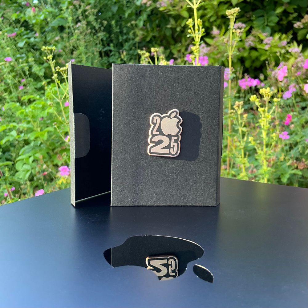 A photo of Apple’s WWDC 2025 pin in its box perched on top of a MacBook out in the garden. 