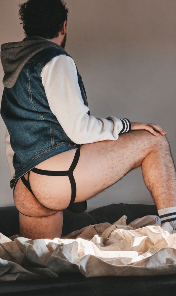 uma foto minha de costas pra câmera, estou vestindo um casaco de moletom com jeans e uma cueca jock preta, ajoelhado em uma perna só, com o outro pé apoiado na frente, mostrando bem minha bunda peludinha 