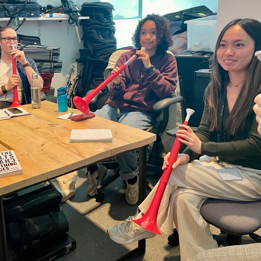 Small group of adult musicians sitting around a wooden table holding and playing red plastic ‘horns’ called PBuzzes. Female in the foreground smiling. 