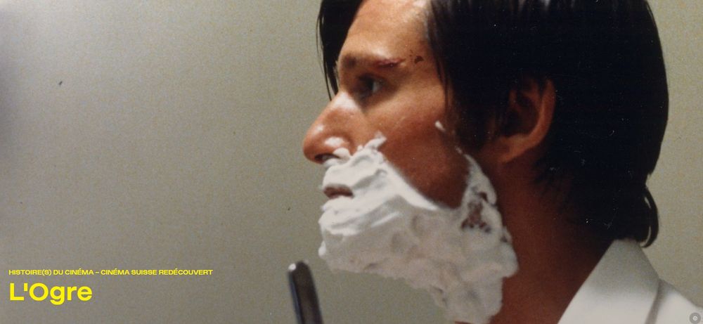 Film still from "L'Ogre": A man, side profile, with dark mid-length hair and shaving foam on his face and a razor in his hand.