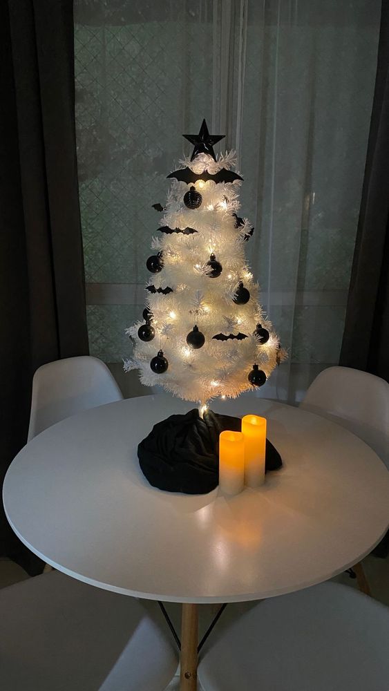 White tabletop Christmas tree, with black bat, and ball ornaments, white lights, and topped with a black star. Sitting on a white table, with two lit white candles nearby.