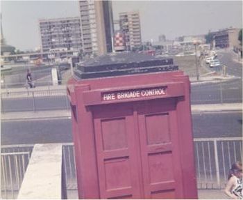 A faded colour photo of a police box painted red, standing beside a wall on a traffic island beside a dual carriageway.