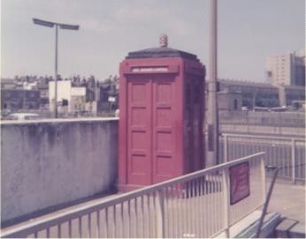 A faded colour photo of a police box painted red, standing beside a wall on a traffic island beside a dual carriageway.
