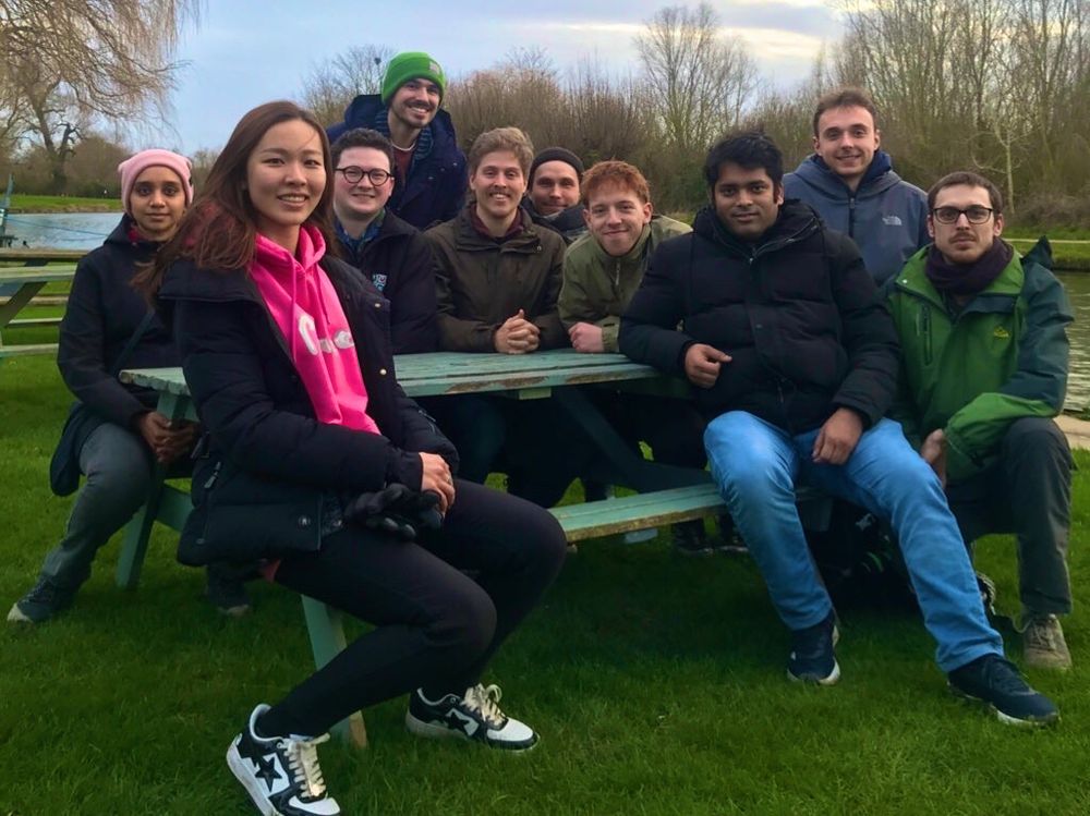A group of 10 sitting on a bench near a river.