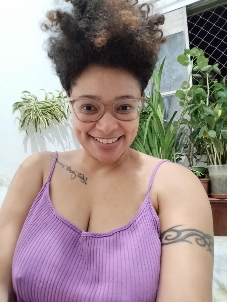 Visto um vestido lilás de alcinha sem sutiã. Bico do seio direito aceso. Sorrio para a câmera. Plantinhas ao fundo e cabelo pra cima.