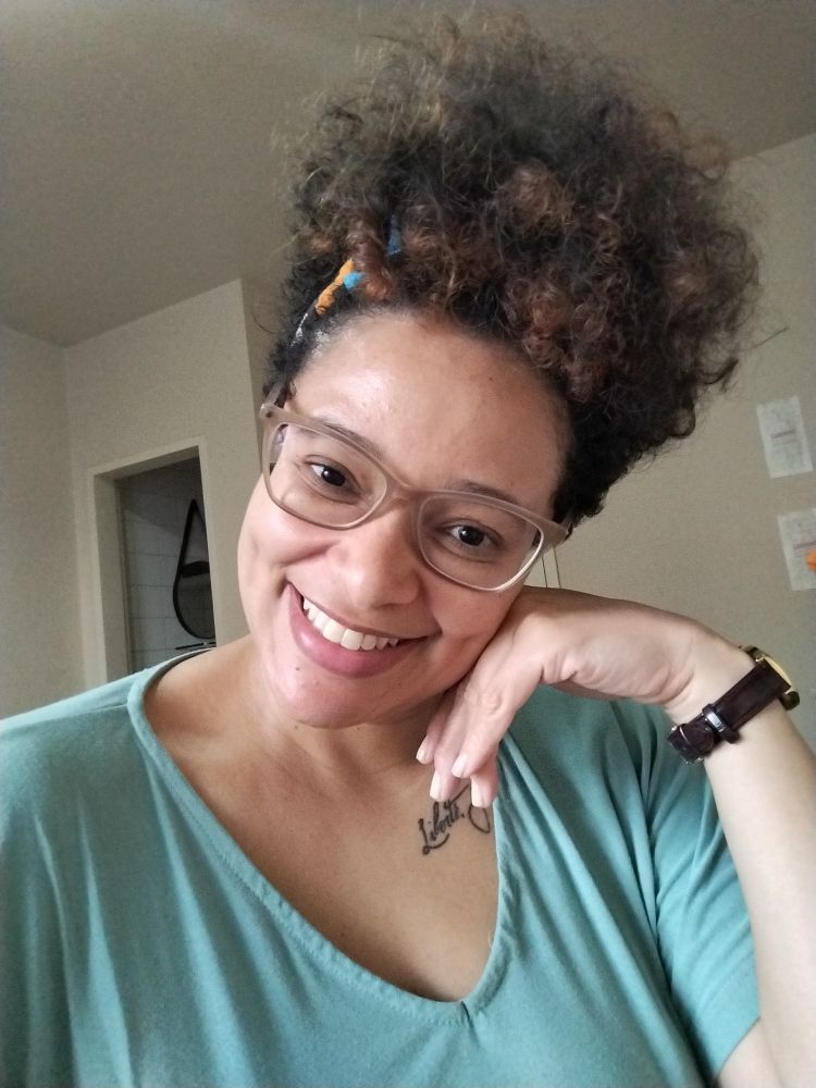 Euzinha muito fofa kkkkkkkk! Macacão verde pistache, cabelos emaranhados pra cima. Pouso o rosto na minha mão esquerda sorrindo com os lábios e os olhos.