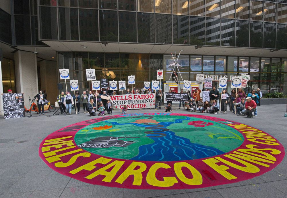 Protesters gather outside Wells Fargo HQ in San Francisco, July 2025. A banner reads Wells Fargo Stop Funding Genocide. A colorful street mural echoes the accusation.
