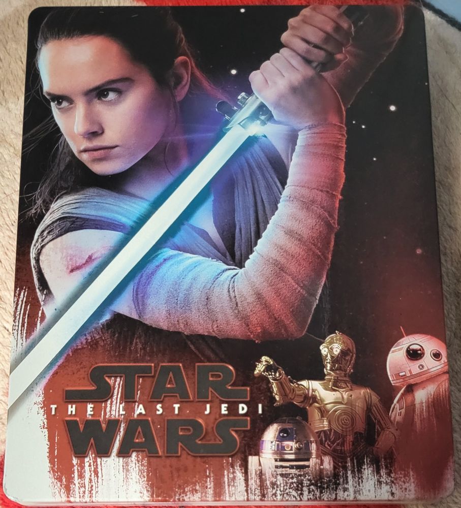 Front cover of the UK steelbook of Star Wars: The Last Jedi. The main colours are red, black and white. Most of the cover shows Rey, who has a determined look on her face and is holding her first/the Skywalker family lightsaber which is ignited. She has a cut on her upper right arm.
On the bottom left is the logo for the movie, it is written as Star Wars with The Last Jedi between Star and Wars. On the bottom right are three droids from left to right they are R2-D2, C-3PO and BB-8.