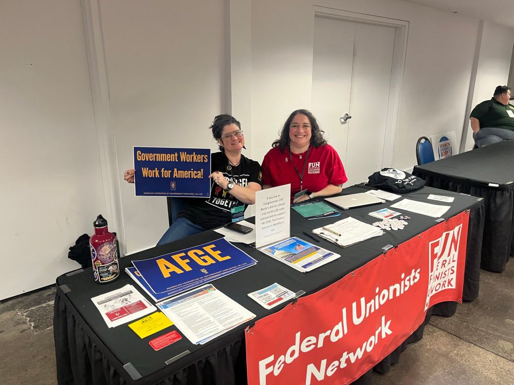 Two dark haired femme presenting people sitting behind an event table, representing the Federal Unionists Network. One is holding an American Federation of Government Employees sign that says "Government Workers Work for America!"