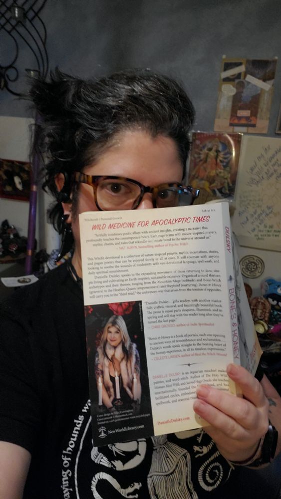 A dark haired person with light skin, wearing glasses and a black T-shirt with Hekate on it. They are partially covering their face with the book "Bones & Honey: A Heathen Prayer Book" by Danielle Dusky. 