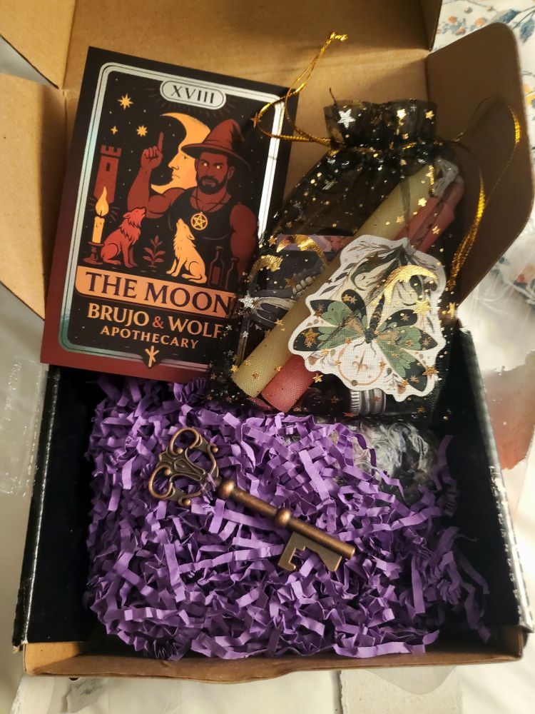 A small cardboard box with shredded purple filler with a bronze decorative key on top, an organza bag with candles, stickers, and ritual oil, and a business card for Brujo & Wolf Apothecary which is modeled after the Moon Tarot Card showing a muscular brujo in a witches hat and wearing a tank top and pentacle necklace. He is holding up one finger. In the foreground are a dark wolf and a light wolf facing each other and howling. Between them is a plant. In the background of the card is a waxing crescent moon man, 3 stars, a castle tower and a lit candle. It's all in shades of copper, gold, silver, and browns. Get yourself some handmade magical goodies!