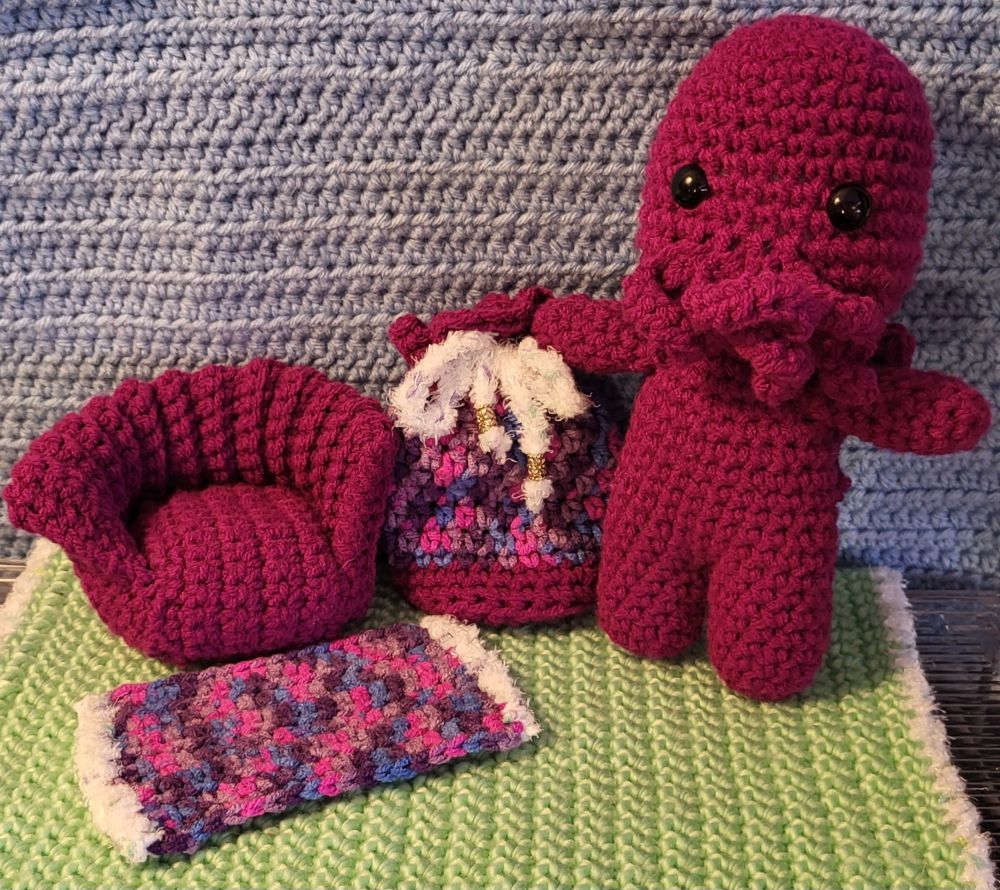 A green ground and blue background of crochet.

A magenta Cthulu (with wings but hard to notice here) stands to the right of a dice bag with similar magenta accents. The chair is also the same magenta. The dice bag and rug are a pink/blue/purple/lavendar mix with puffy white fringe.
