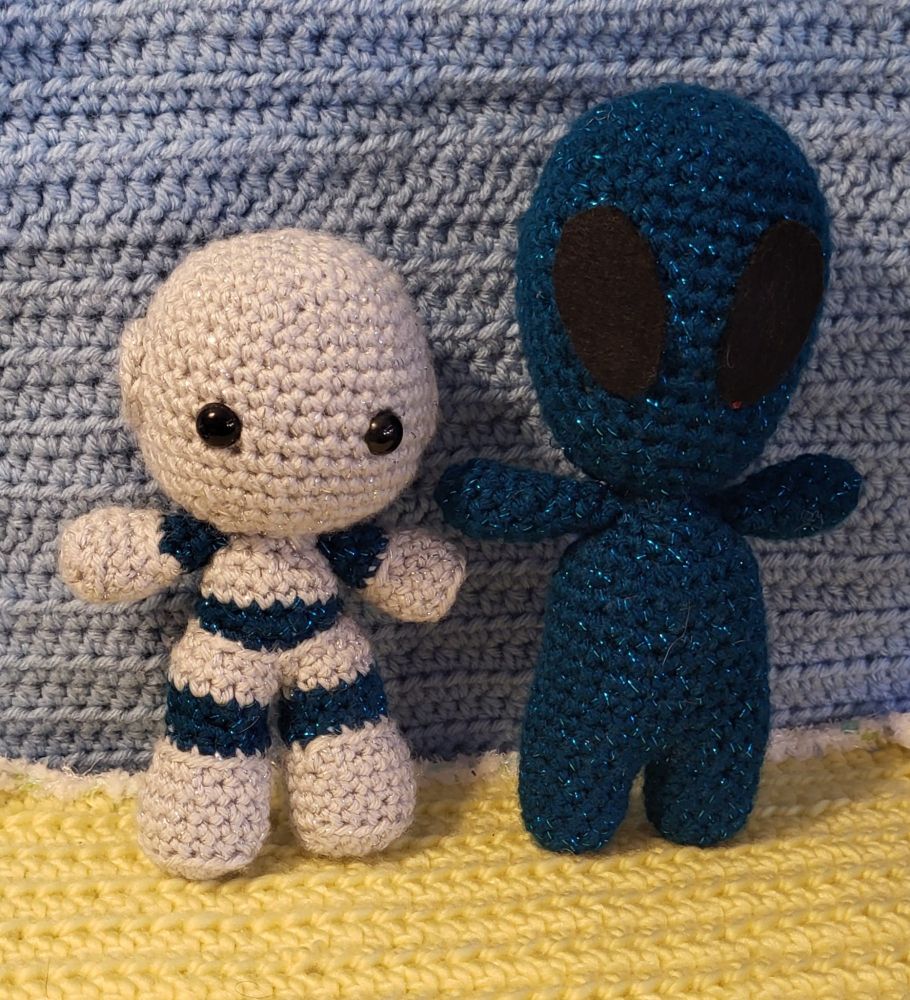 A crochet background of yellow ground and blue wall. To the left, a grey robot with blue glitter joints at the arm, legs, and torso. He has a bolt on each side of the head where ears would be. To the right, and entirely blue glitter alien with large oval cut eyes on an oversized oval head.