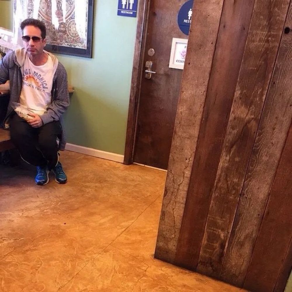 A photo of David Duchovny sitting next to a unisex restroom. He looks a bit sad as he holds a bag of what might be granola. 
