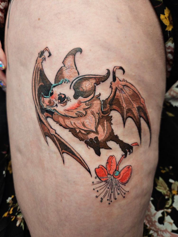 A tattoo of a fruit bat on the back outer side of a person's thigh. The bat is smiling holding a guava blossom, and the lines are illustrative and show movement. The bat is colored in shades of warm brown with pink cheeks and nose, while the flower is hot pink. There are a few highlights in seafoam green that compliment nicely.
