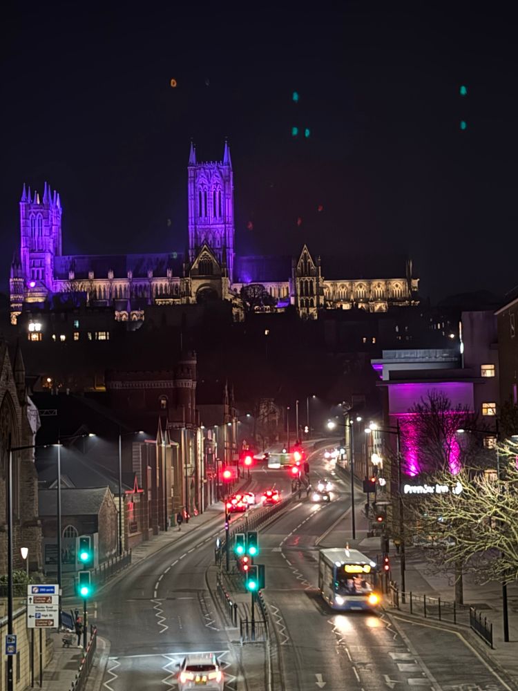 A nighttime view over a Lincoln city street towards Lincoln cathedral on the hill. The cathedral towers are illuminated in purple light for Lent. 