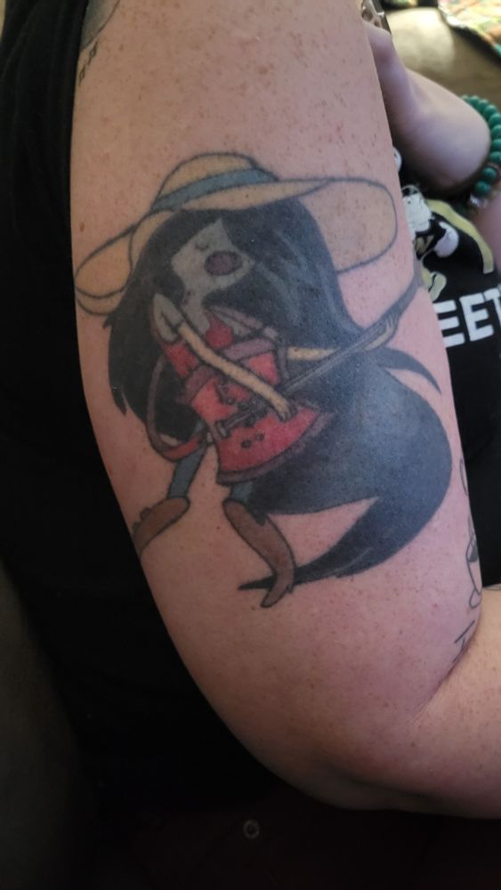 My upper arm with a tattoo of Marcilene the Vampire Queen from the episode What Was Missing. A slender vampire, grey in color wearing blue pants, knee high brown boots a red tank top and a large yellow sun hat with a blue ribbon around the inside brim. She has a ton of black hair that falls to her feet. She is holding a red bass guitar and looks to be singing (spoiler alert:she most definitely is singing)