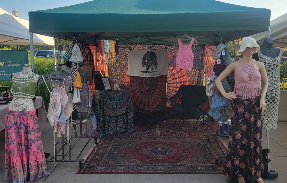 green Vendor tent with a mannequin on either side wearing boho skirts and crocheted crop tops in green and pink respectively. 70's style red rugs with similar mandala tapestries in black and orange hanging from behind. Pink crocheted tank top hanging from the front. Rack on either side with crocheted clothing hanging from them. A sign in the middle that reads Willowmecreations and has a weeping willow tree with a ball of yarn in a swing from its branches. 