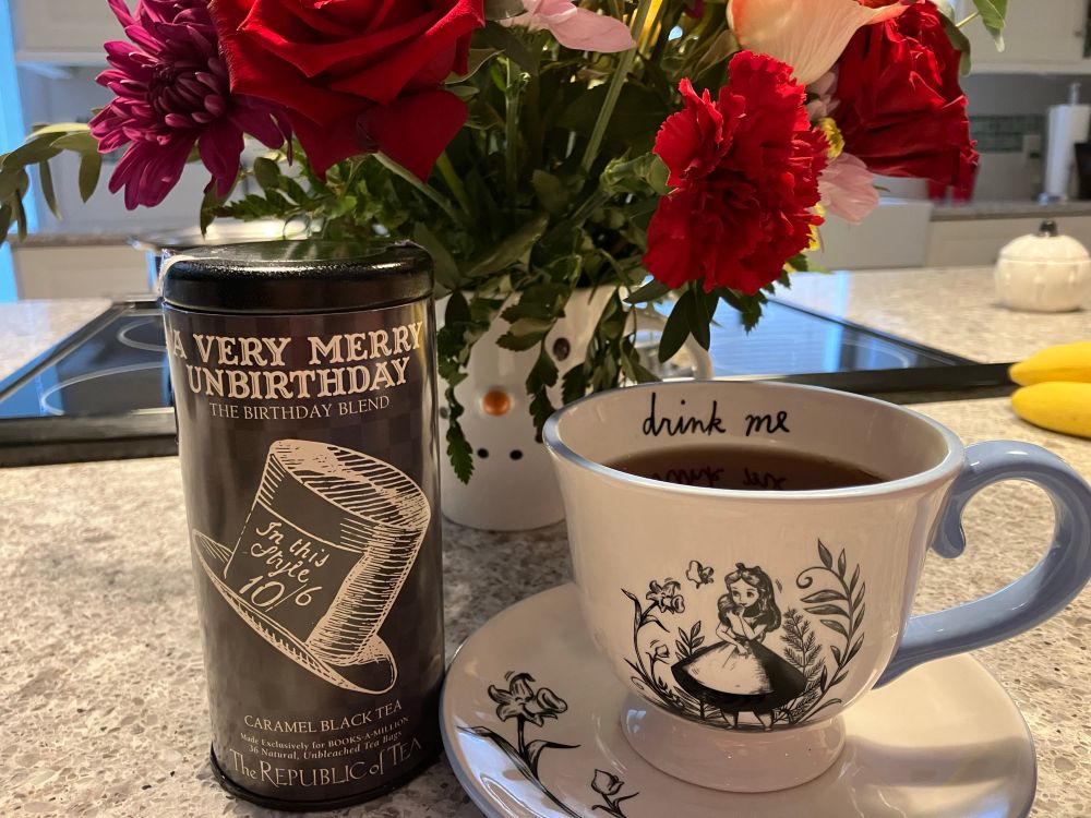 A cup of tea in an Alice in Wonderland teacup and saucer, next to “A Very Merry Unbirthday” caramel black tea, in front of flowers.